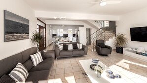 A spacious living area with a sofa set, a coffee table with decorative items, a TV mounted on the wall, and a staircase leading to an upper level. - Faros Seaside Villa Larnaca (Pervolia)