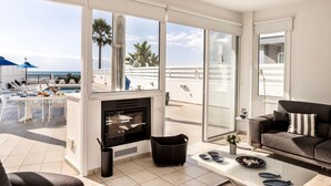 A modern living room with a fireplace, a sofa, and a coffee table. There is a balcony with a pool and palm trees visible through large windows. - Faros Seaside Villa Larnaca (Pervolia)