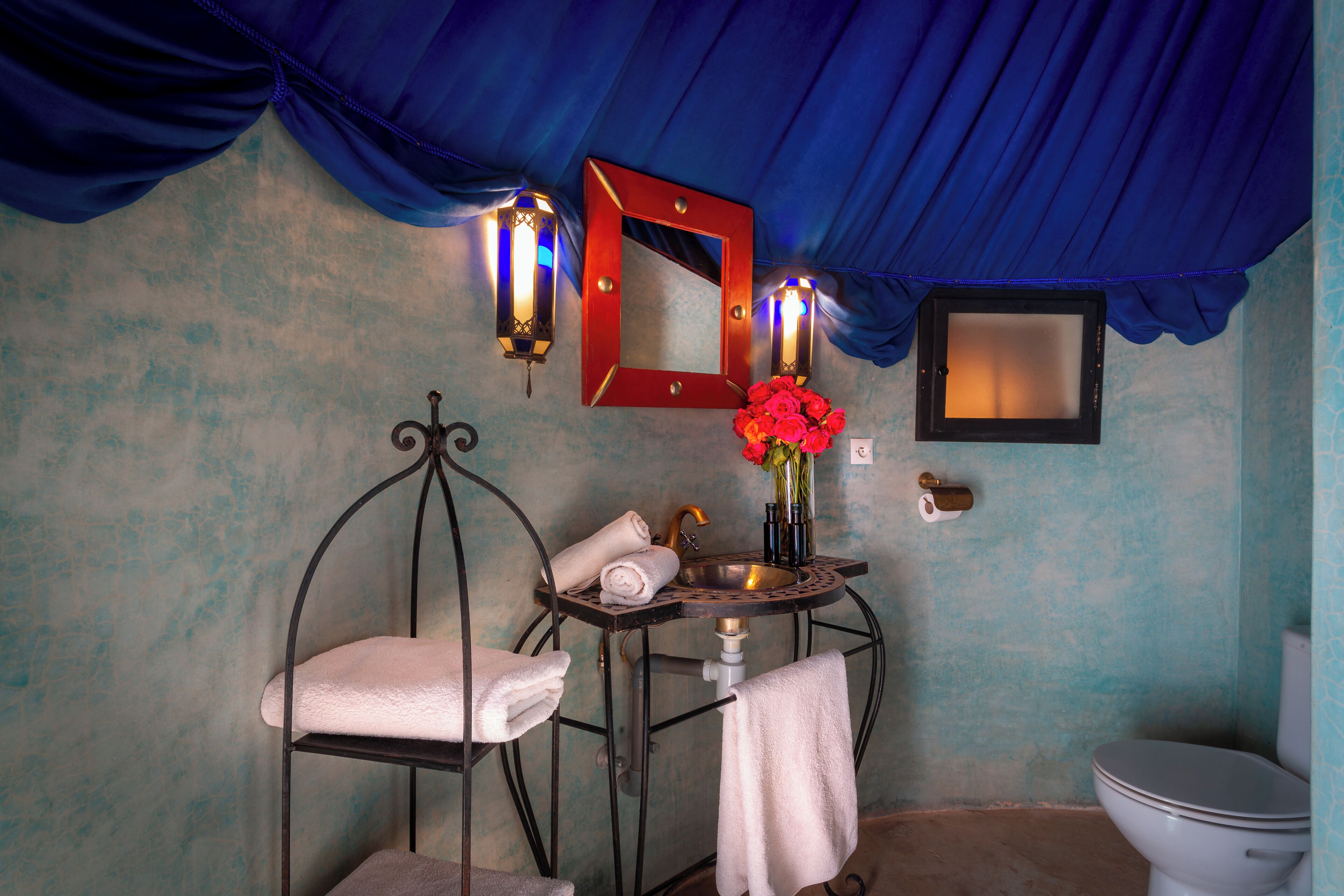 Un baño con techo azul, un lavabo con espejo, un florero con flores rojas y un inodoro.