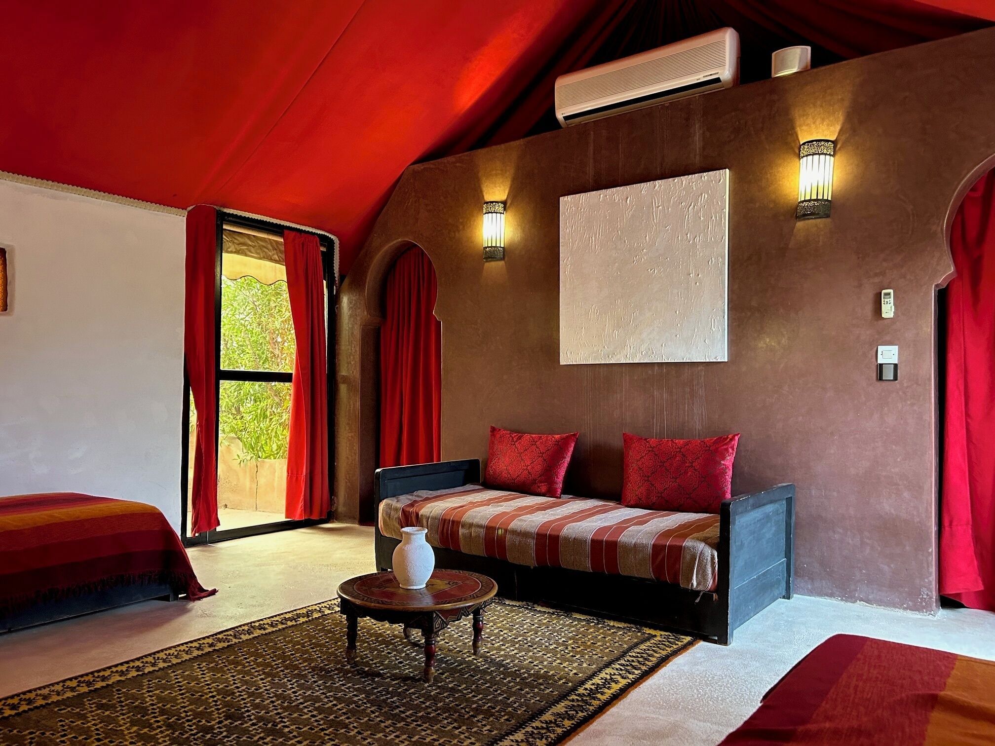 A room with a sofa, a small table with a vase, a patterned rug, and a large window with red curtains.