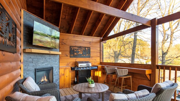 Une charmante cabane en rondins avec une cheminée, un coin salon extérieur et un téléviseur fixé au mur.