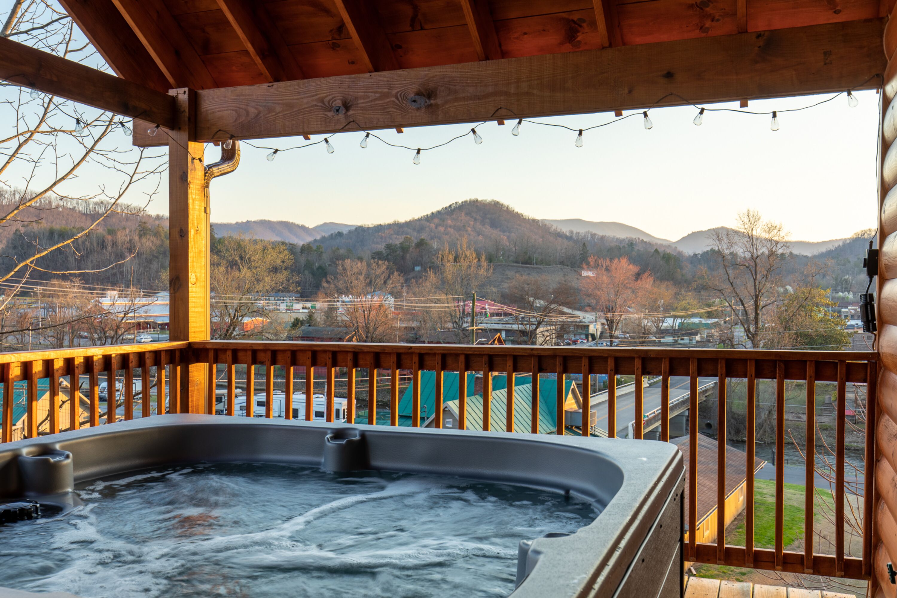 A hot tub under a wooden pergola with a view of mountains and a town.