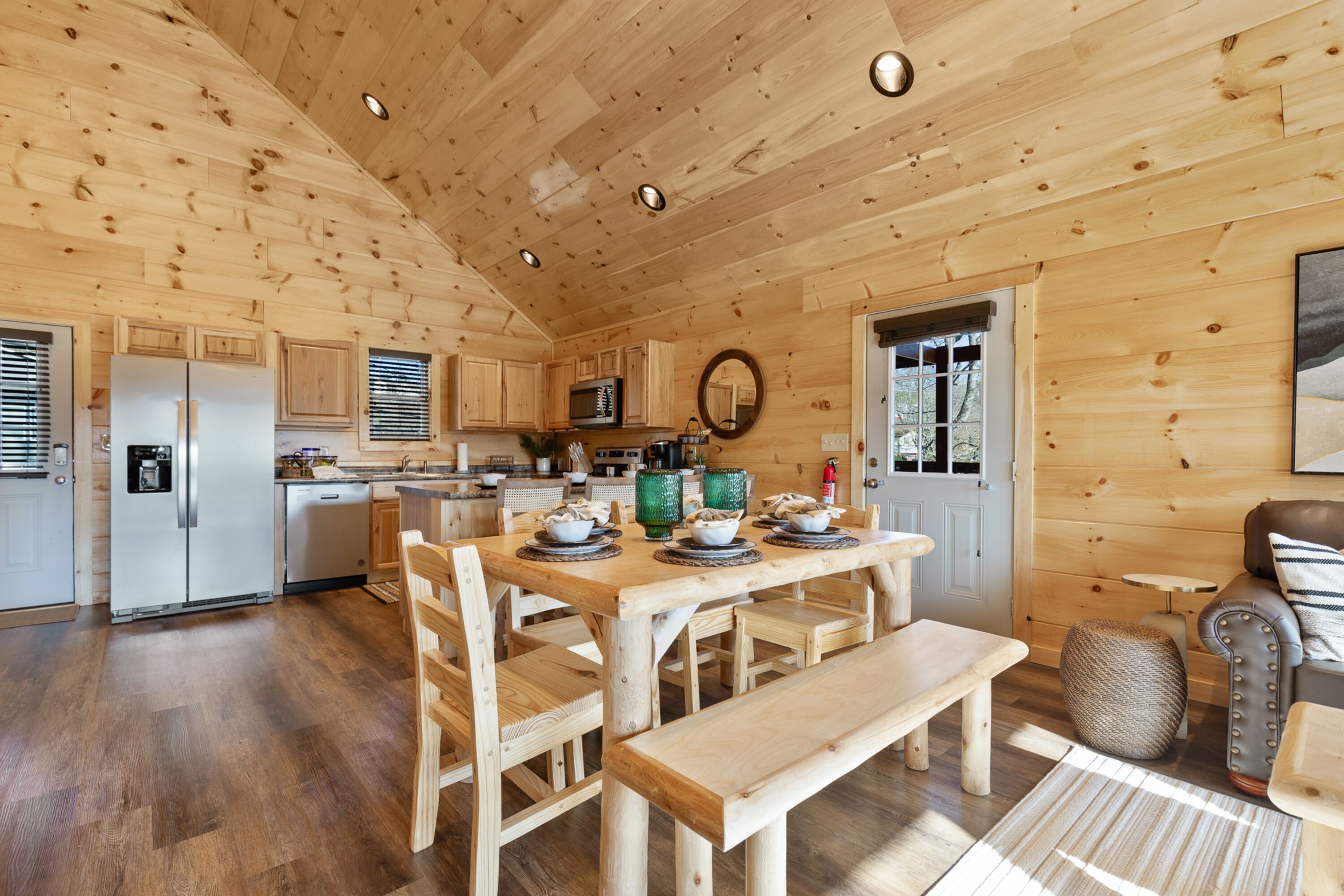 Interior de una cabaña de madera con mesa de comedor, sillas, banco, sofá, mesita auxiliar y una zona de cocina con electrodomésticos.