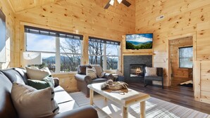 L’intérieur chaleureux d’un chalet avec une cheminée, des murs en bois, un canapé et de grandes fenêtres offrant une vue panoramique.