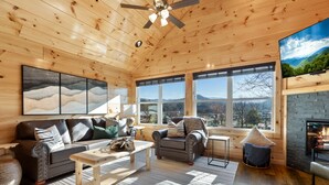 L’intérieur chaleureux d’un chalet avec un plafond en bois, une cheminée, un canapé, un fauteuil et une grande fenêtre donnant sur une vue imprenable sur les montagnes.