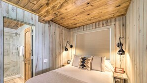 A bedroom with a wooden ceiling, a large bed with a white bedspread, two bedside lamps, a window with blinds, and a wooden nightstand with a picture frame.