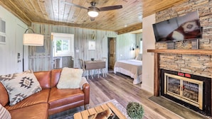 A cozy cabin interior with a wooden ceiling, a fireplace, a television mounted above it, a dining area with a table and chairs, and a bed in the background.