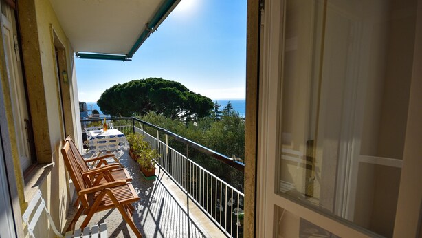 A balcony with wooden chairs and a table, overlooking a coastal area with trees and the sea. - Relax Apartment N 5 by Wonderful Italy (Sanremo)