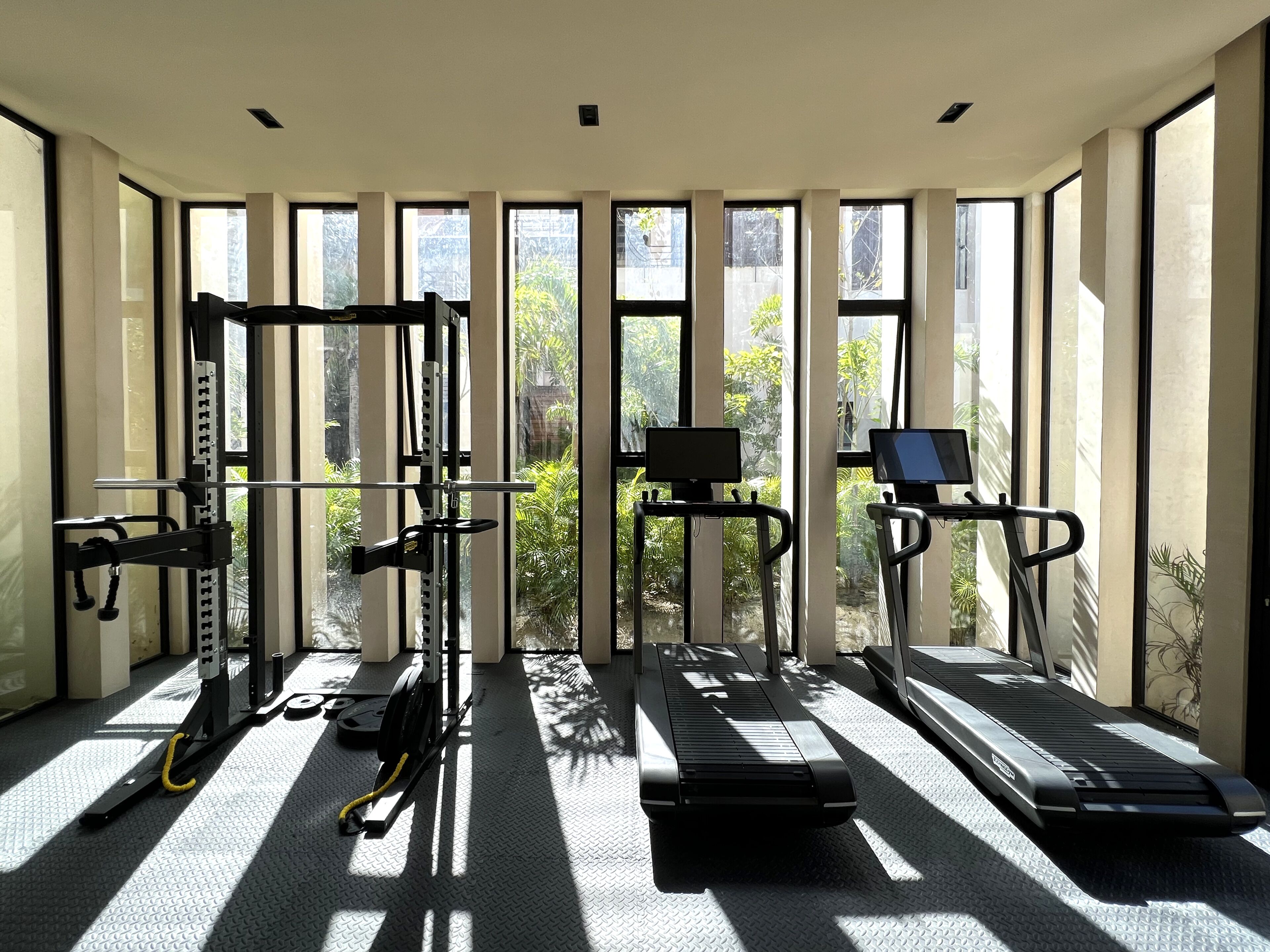 A gym with treadmills, a weight machine, and a stationary bike, with large windows and a view of greenery outside.