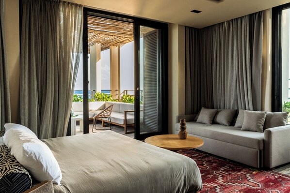 A modern hotel room with a bed, sofa, and a small round table. There is a balcony with a view of the sea.