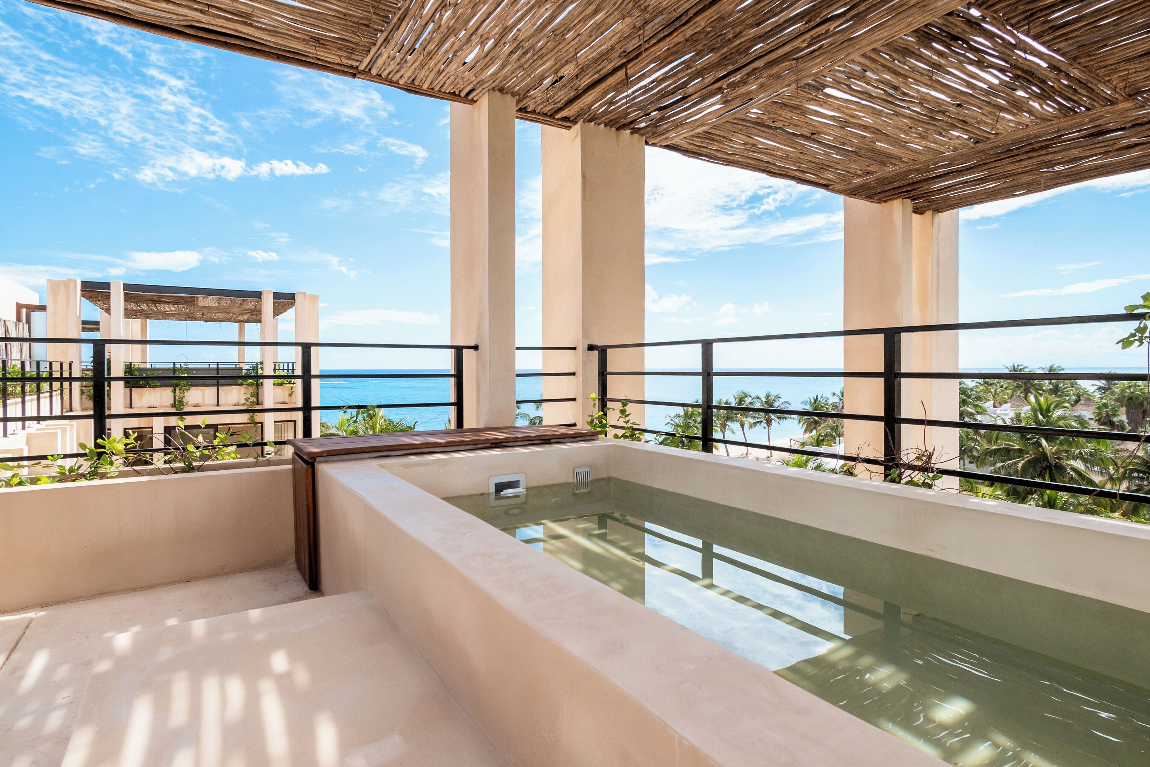 A rooftop pool with a view of the ocean, surrounded by palm trees and a thatched roof structure.