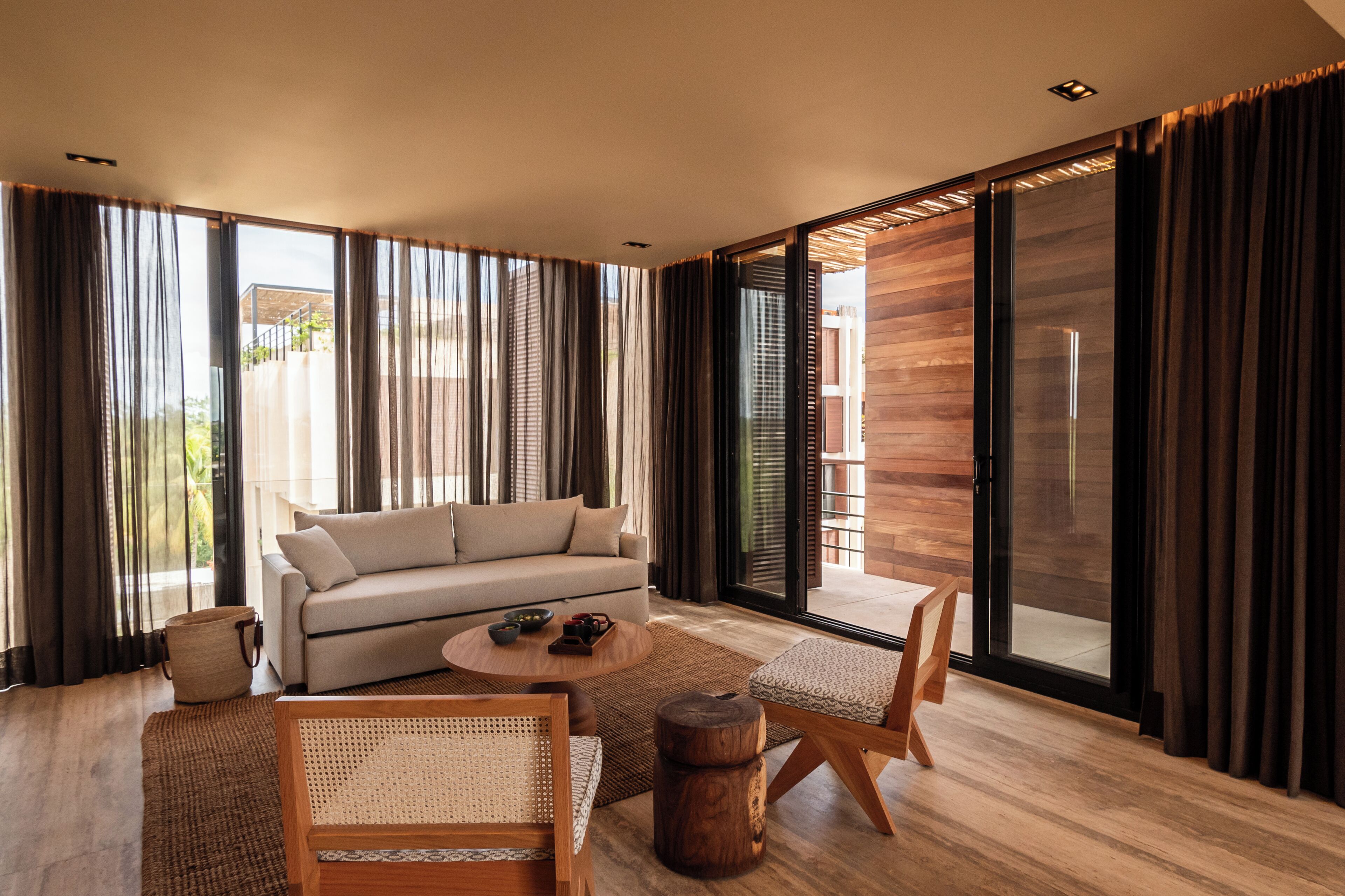 A modern living room with a sofa, coffee table, and chairs, large windows with curtains, and a wooden floor.
