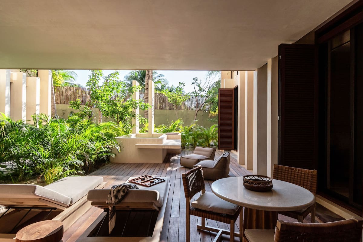 A sunlit patio with wooden furniture, a round table, and lush greenery.