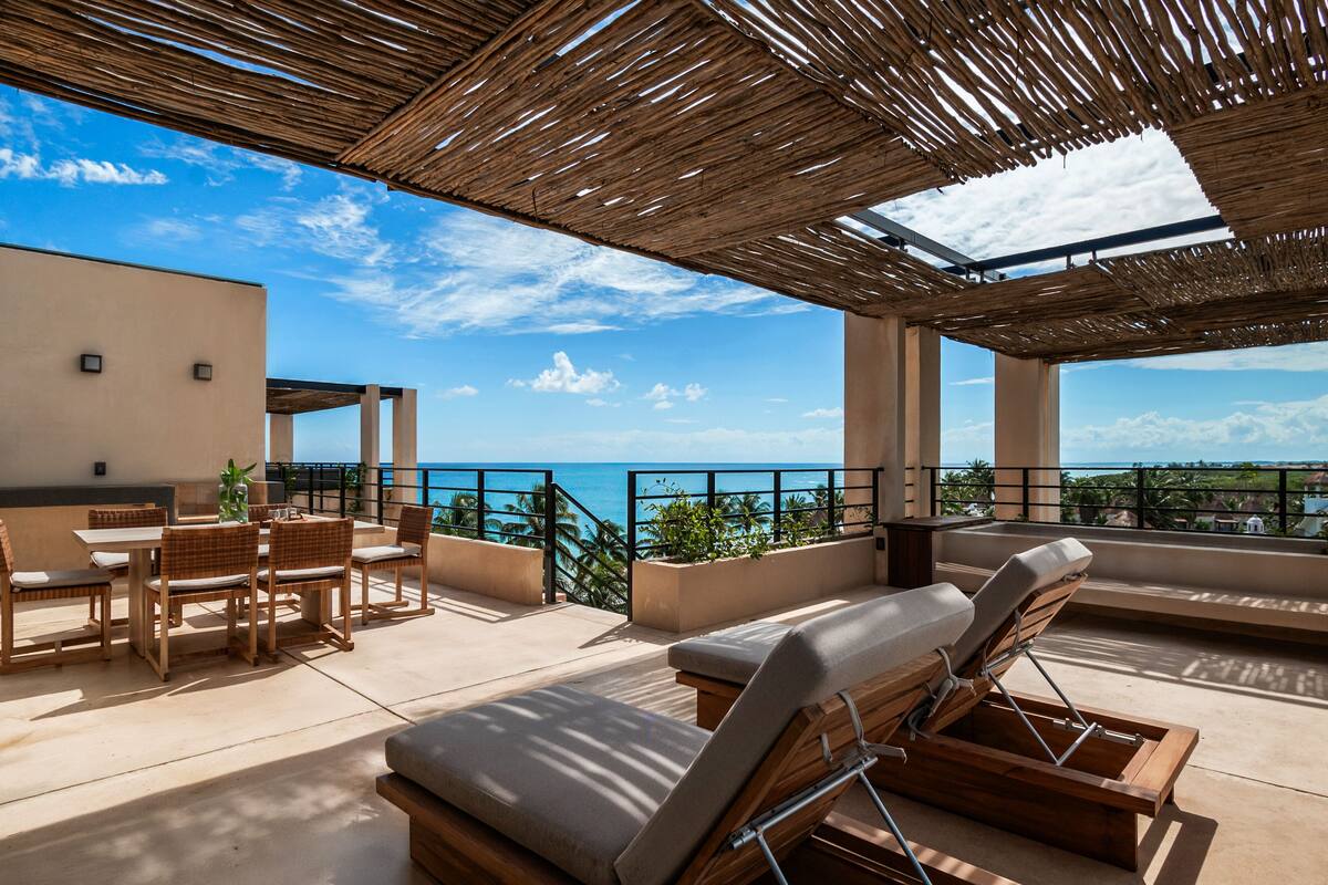 A rooftop terrace with a view of the ocean, wooden furniture, and a thatched roof.