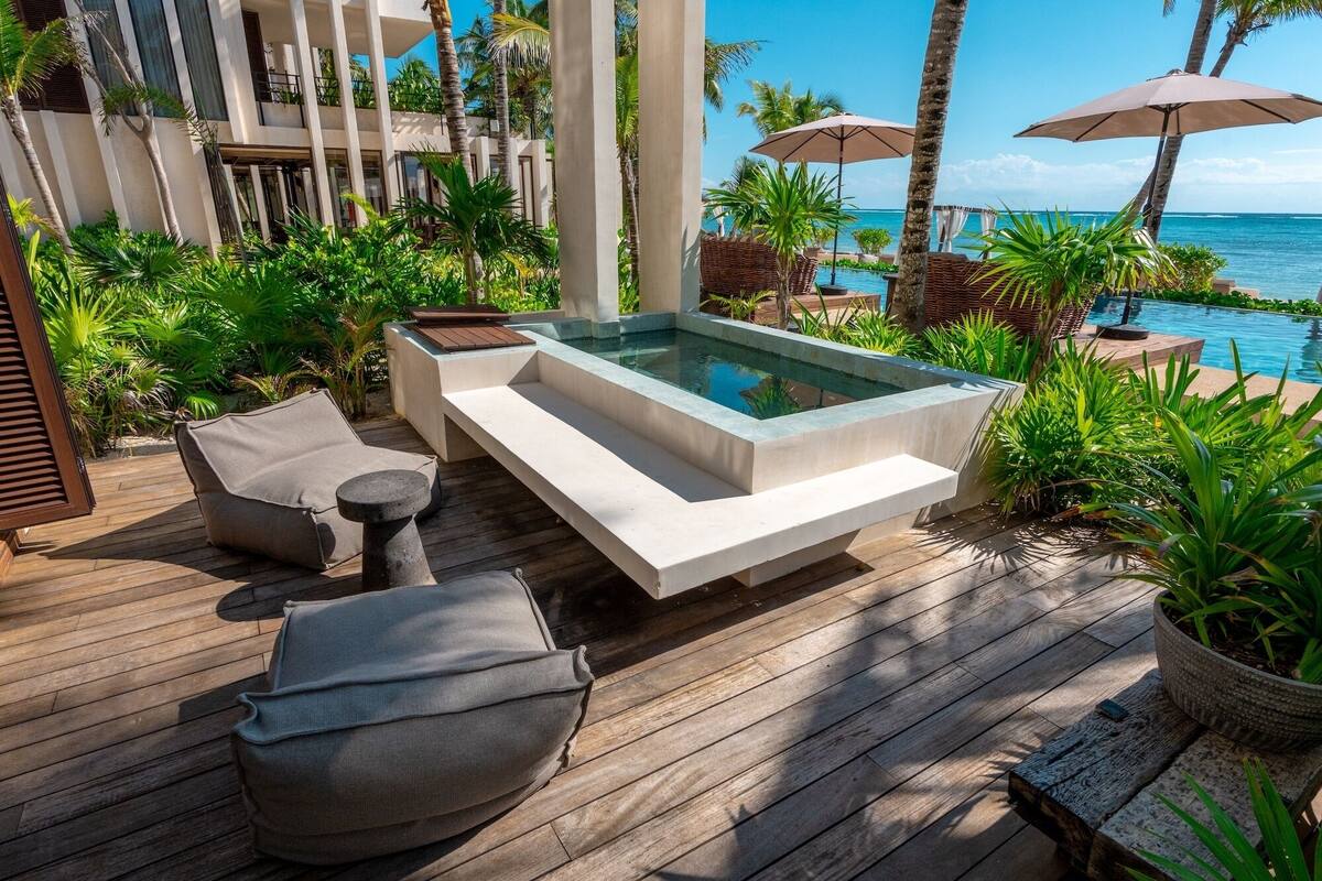 A wooden deck with a hot tub, lounge chairs, and umbrellas overlooking a pool and ocean.