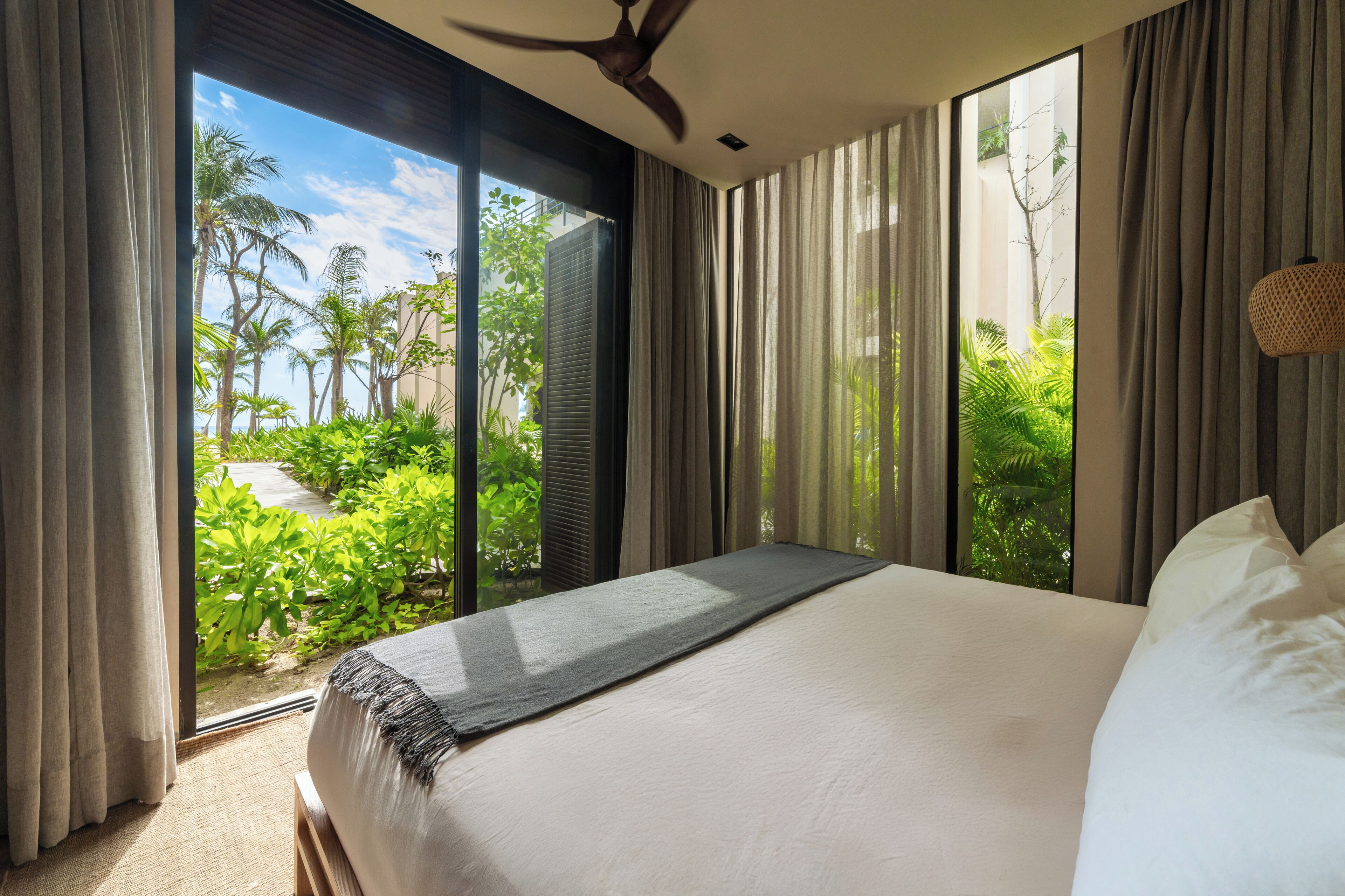 A bedroom with a large bed, a ceiling fan, and a view of lush greenery outside.