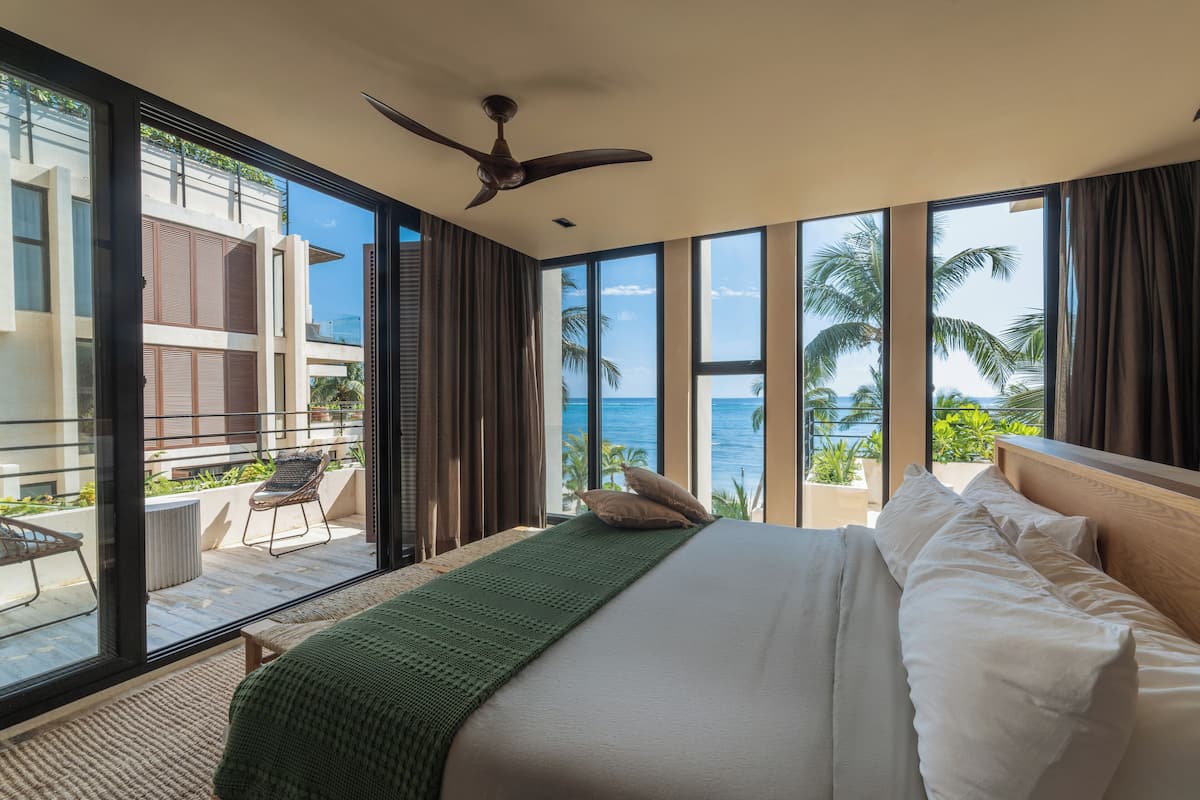 A bedroom with a large bed, a ceiling fan, and a view of the ocean through large windows.