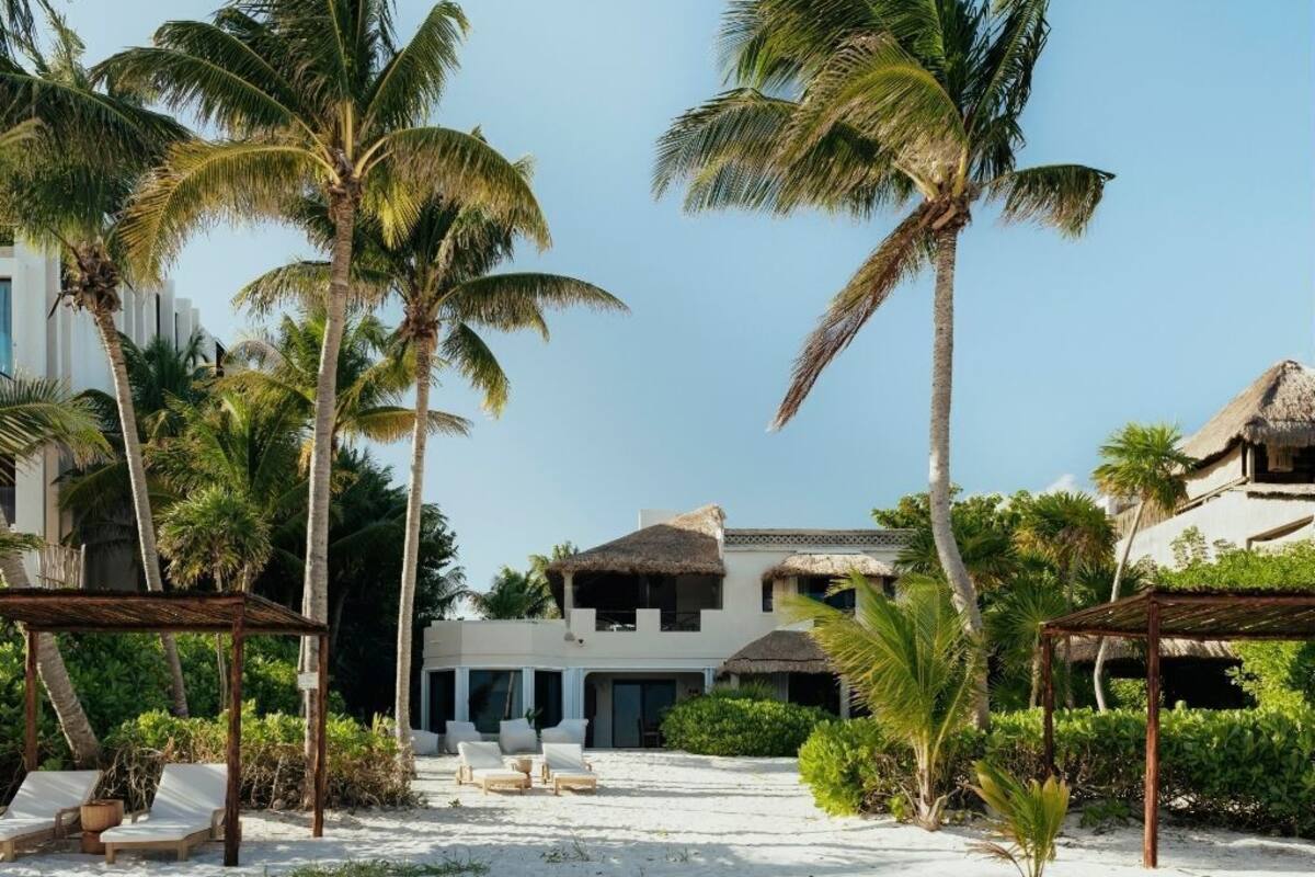 A beach resort with thatched-roof buildings, palm trees, and a sandy beach with lounge chairs.