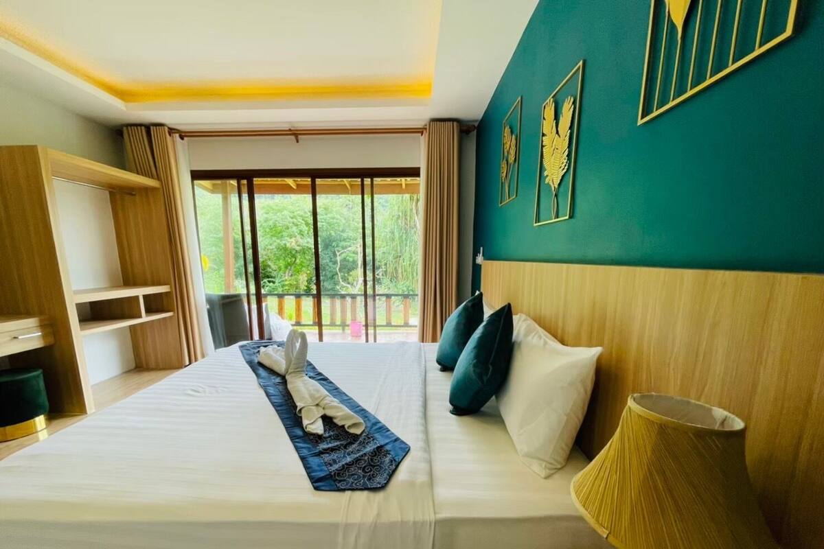 A hotel room with a large bed, a bedside lamp, and a view of greenery through the window.