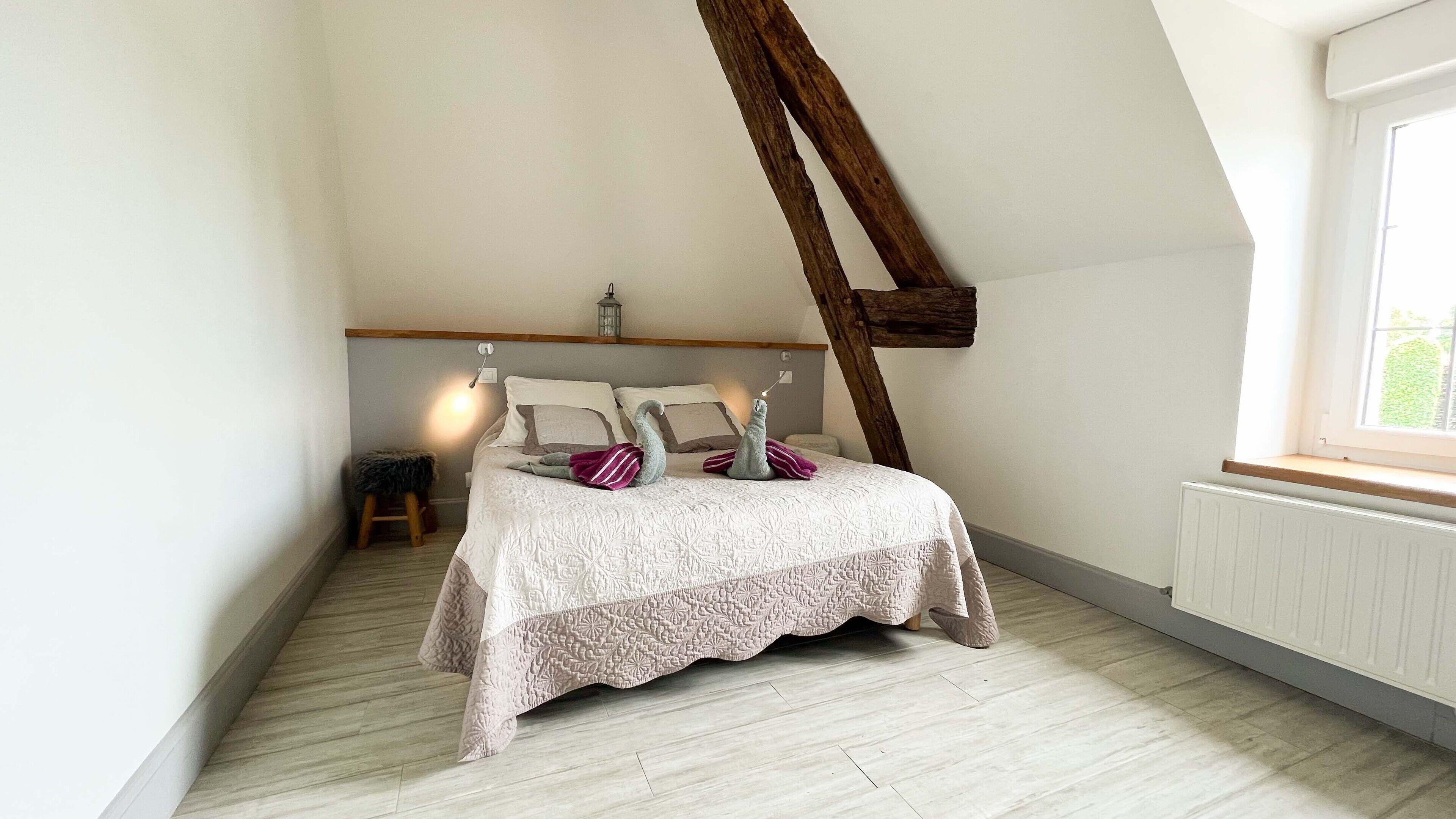 Ein Schlafzimmer mit schrägem Dach, einem Bett mit weißer Bettdecke, Kissen und einem kleinen Hocker.