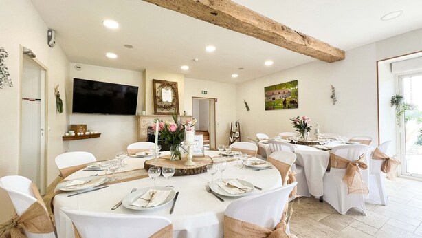 Een eetgedeelte met ronde tafels, witte tafelkleden en stoelen met beige linten. Op de tafels staan bloemige decoraties.
