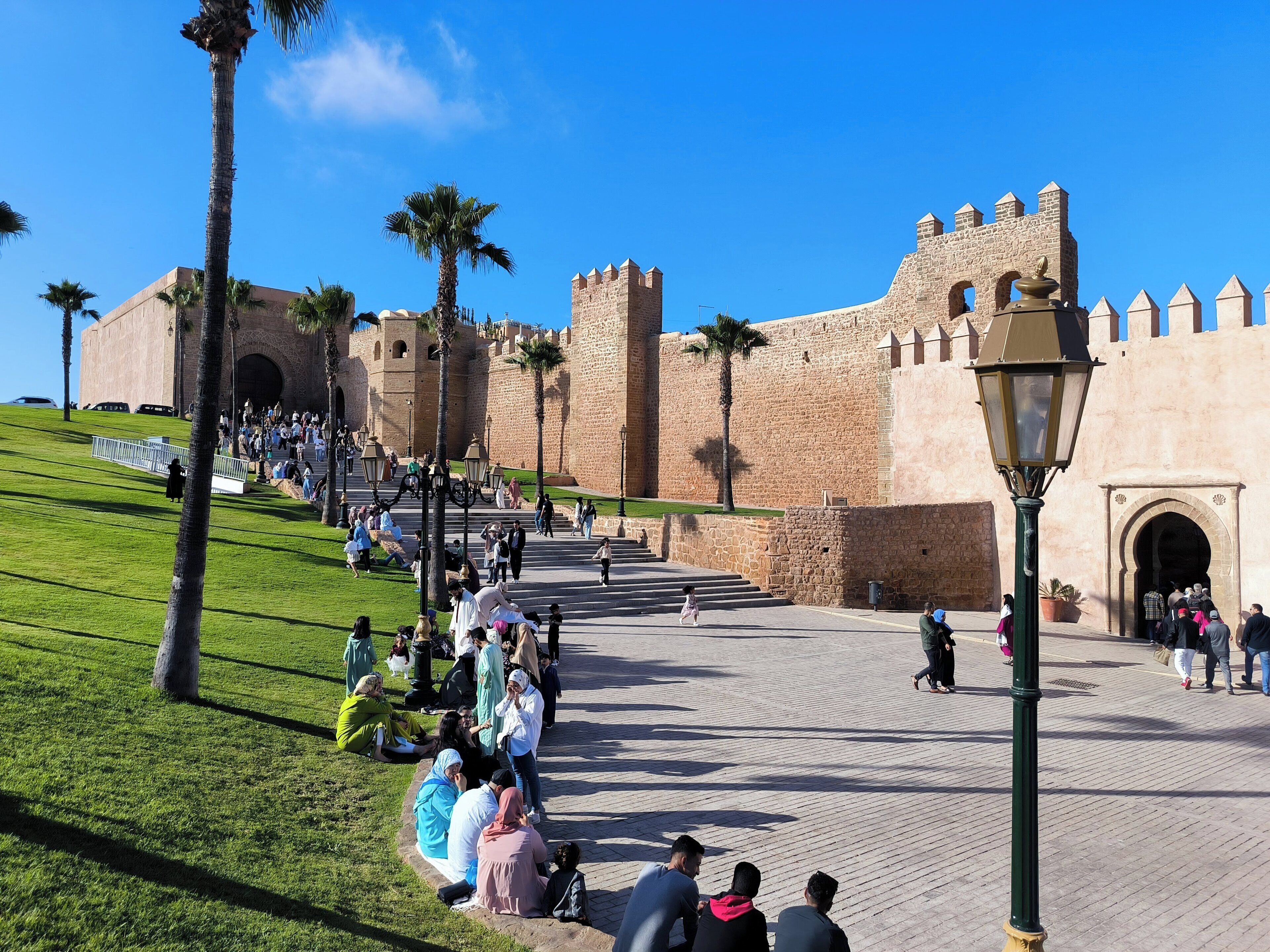 Una fortaleza histórica con muros de piedra, palmeras y una plaza pavimentada con gente sentada y caminando.