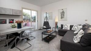 Una sala de estar moderna con un sofá, una mesa de centro de vidrio y una silla. Hay una ventana con vistas a árboles y una puerta que conduce a otra habitación.