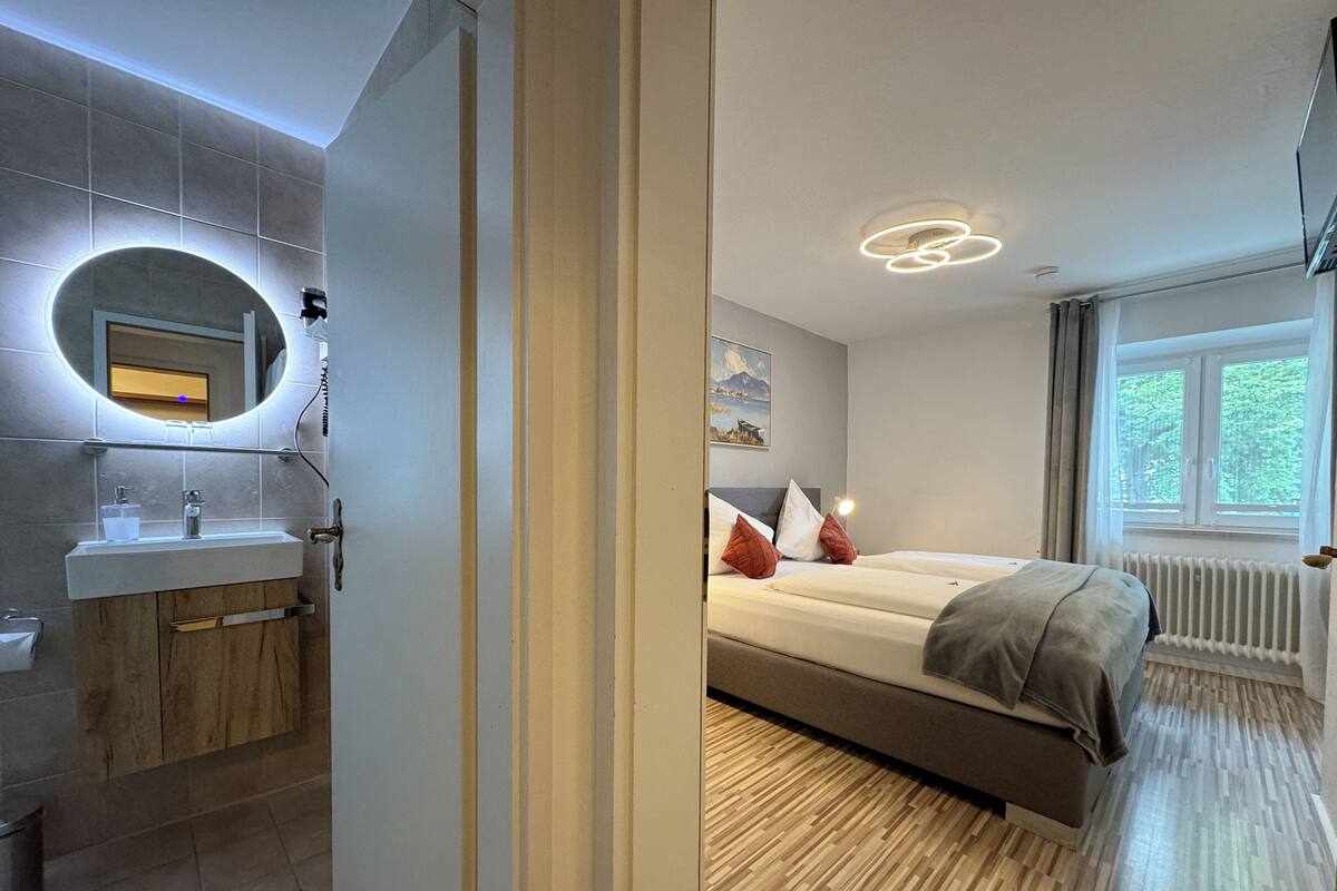 A modern hotel room with a wooden floor, a large bed with a headboard, a bedside table, a mirror with a light, a window with a view of greenery, and a bathroom with a sink and mirror.