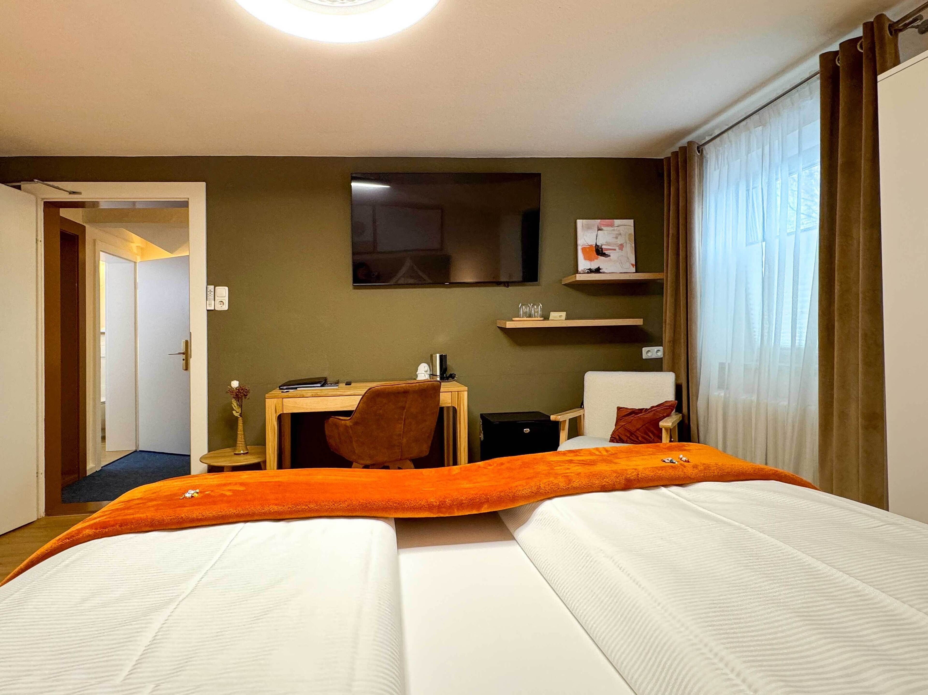 A hotel room with a bed, a desk, a chair, a small table, a mirror, and a window with curtains.