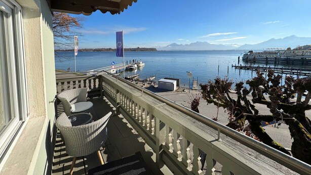 A balcony with wicker furniture, a view of a calm lake, a dock with boats, and a mountain in the distance. - Gästehaus Sieben Zimmer am See  (Prien am Chiemsee)