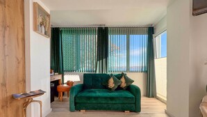 A modern living room with a green sofa, wooden floor, and a view of the outdoors through large windows. - Gästehaus Sieben Zimmer am See  (Prien am Chiemsee)