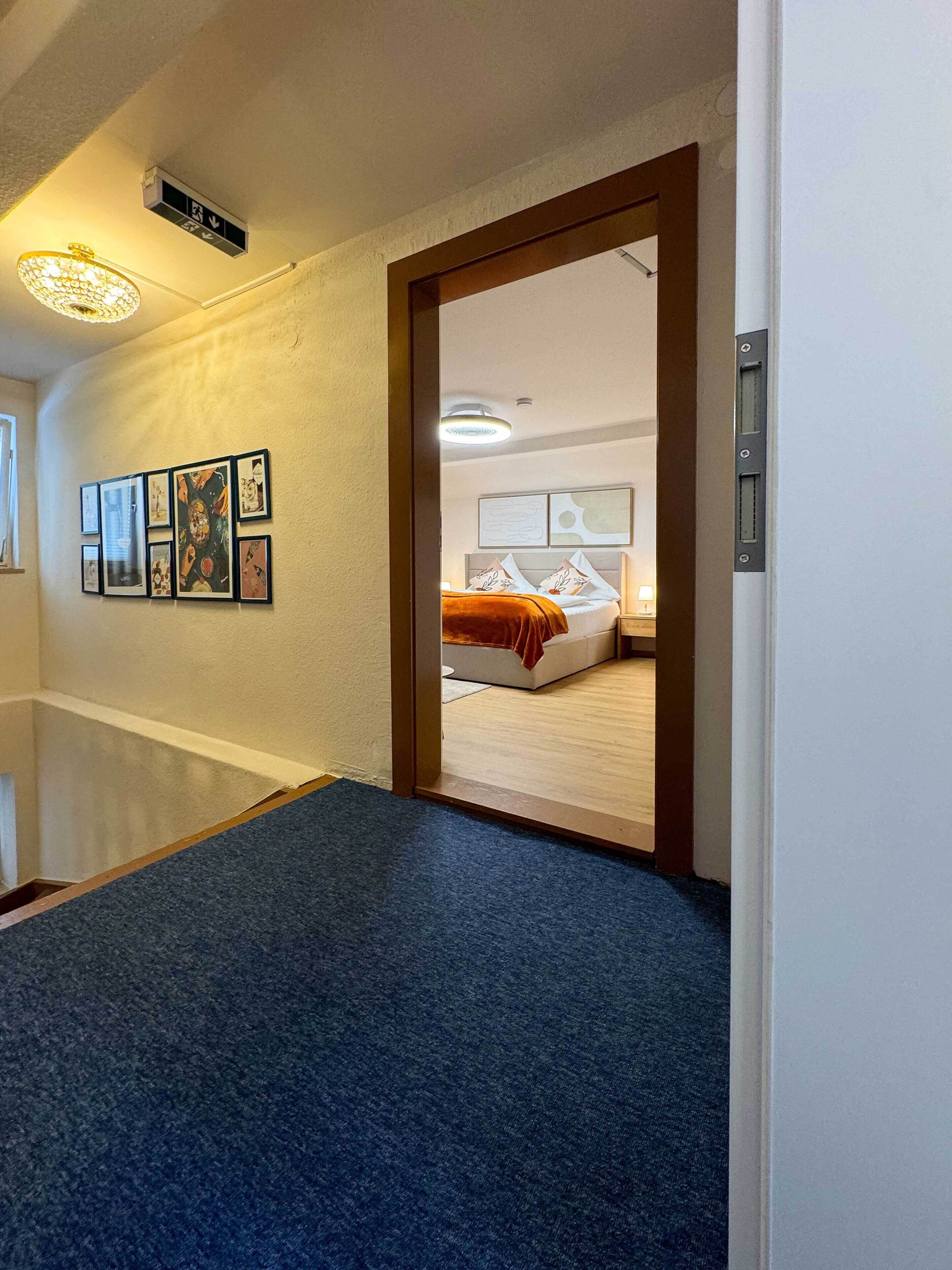 Pasillo de hotel con alfombra azul que conduce a un dormitorio con cama, mesita de noche y lámpara de pared. En la pared hay cuadros enmarcados y una luz en el techo.