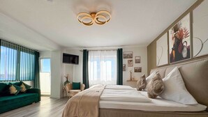 A modern bedroom with a large bed, a green sofa, a TV, and a view of the city. - Gästehaus Sieben Zimmer am See  (Prien am Chiemsee)