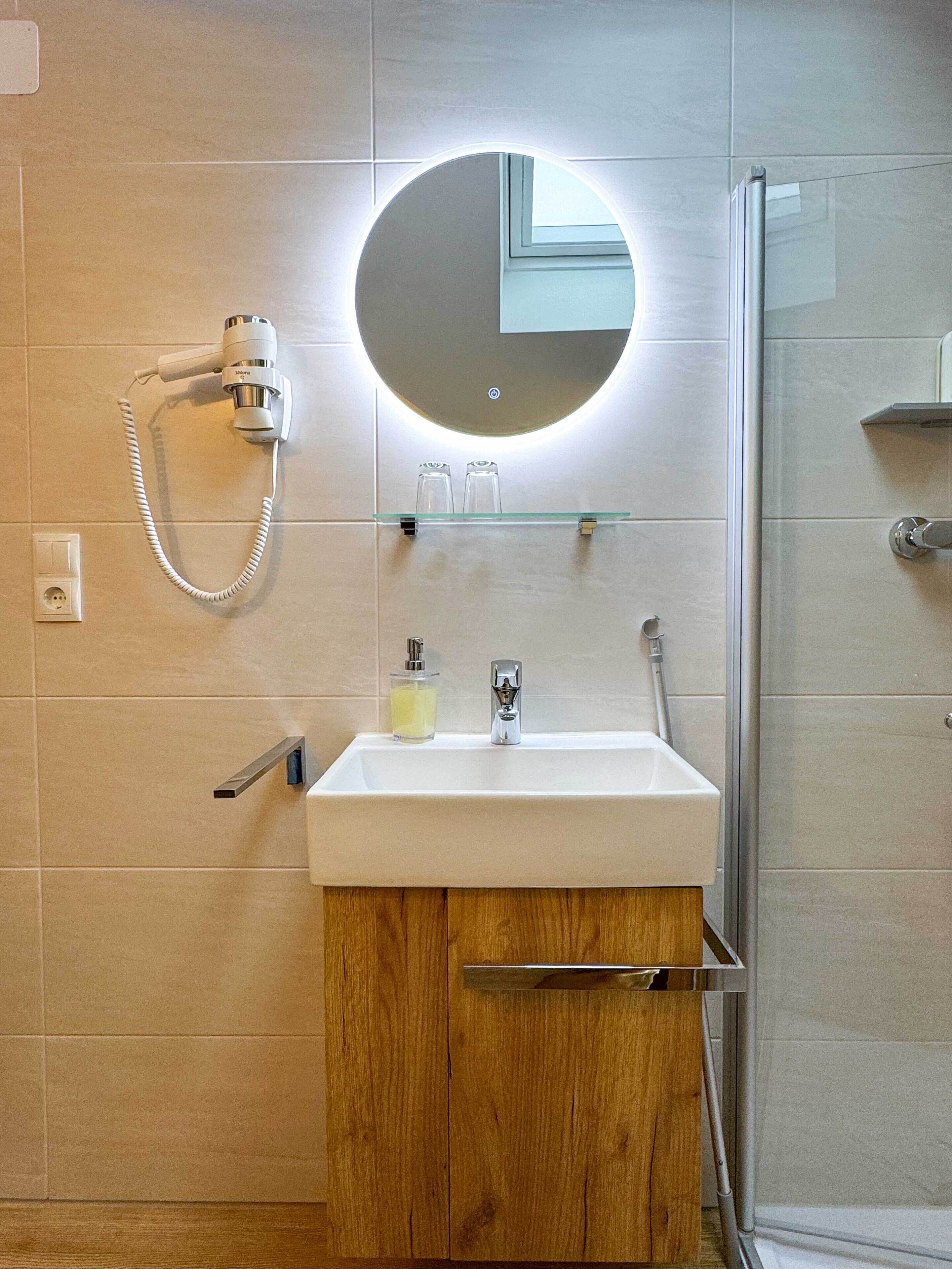 Ein Badezimmer mit weißem Waschbecken, einem Spiegel, einem Föhn und einer Duschkabine aus Glas.