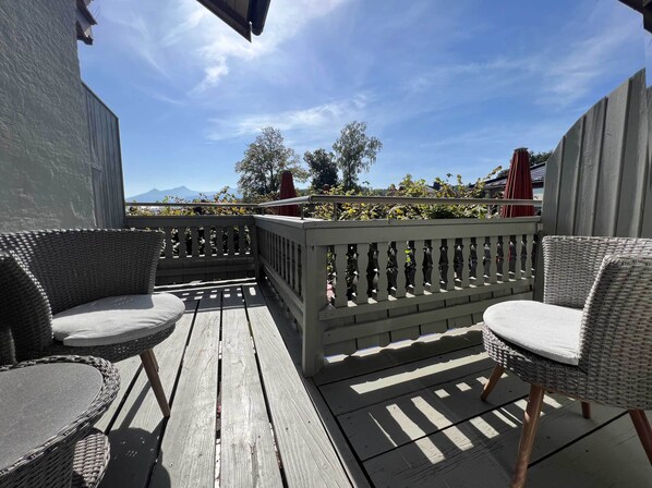 A wooden deck with wicker chairs and a table, overlooking a garden and mountains. - Gästehaus Sieben Zimmer am See (Prien am Chiemsee)