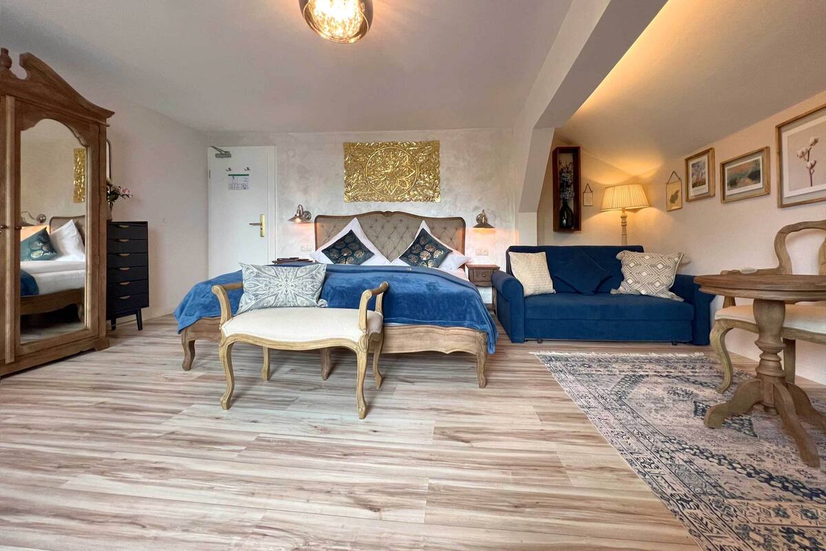 A bedroom with a large bed, a blue sofa, a wooden chair, a small table, and a decorative wall piece.