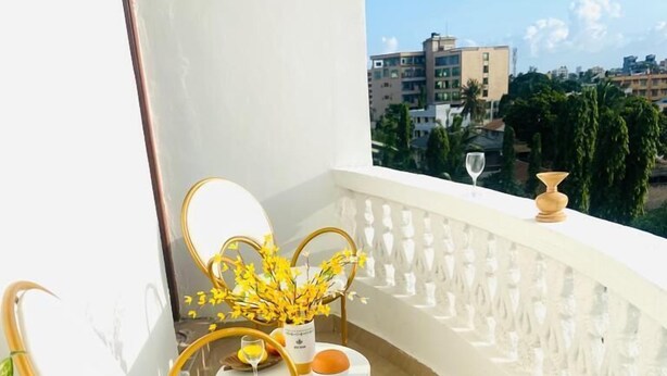 A balcony with two white cushioned chairs and a small table, a vase with yellow flowers, and a glass of orange juice. - Lux Suites Bamburi Cozy Apartments (Mombasa)