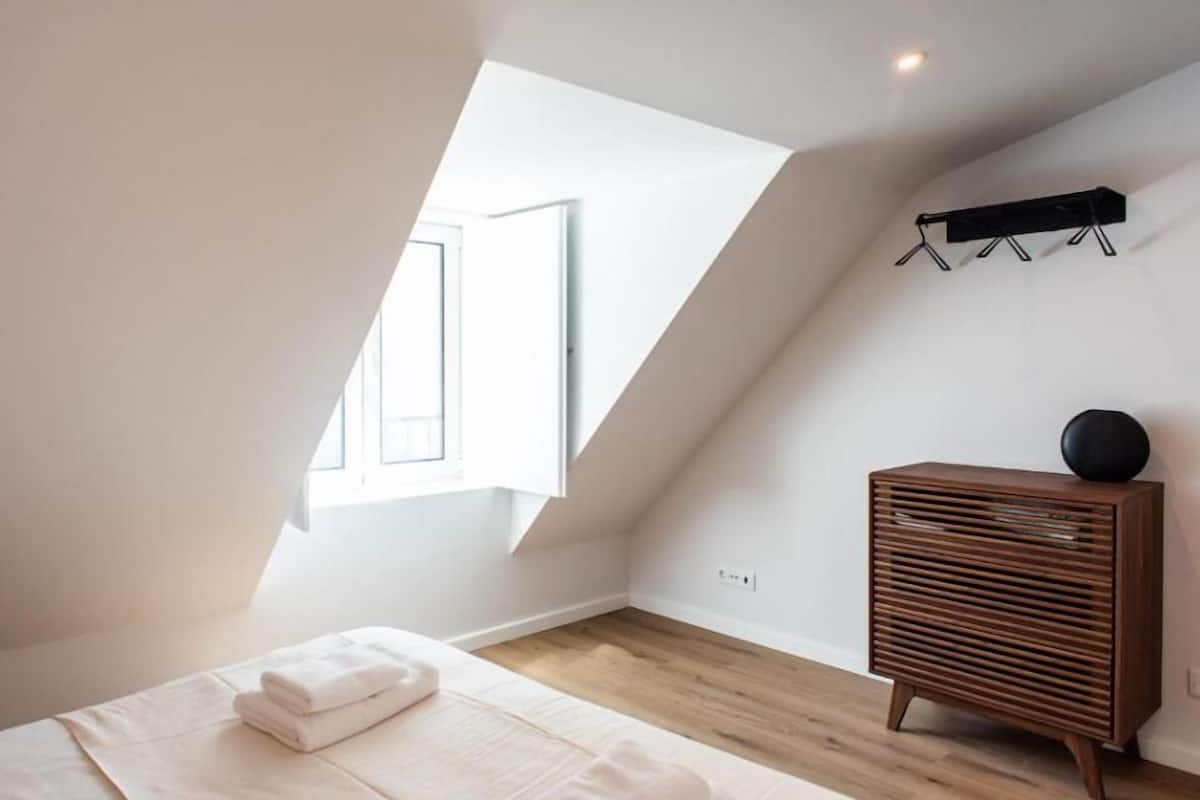 Un dormitorio con techo inclinado, un aparador de madera, una mesita de noche con lámpara y una ventana que proporciona luz natural.