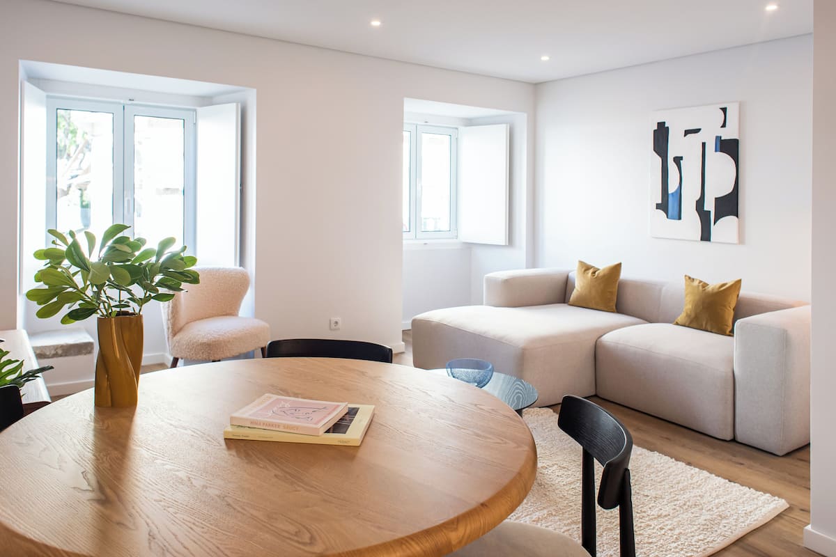 Un salón moderno con una mesa redonda de madera, un sofá color beige y una silla. Hay una planta en maceta sobre la mesa y una ventana con marcos blancos.