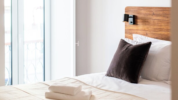 Een hotelkamer met een bed, een houten hoofdbord, een wit kussen, een donker kussen en een raam met uitzicht.