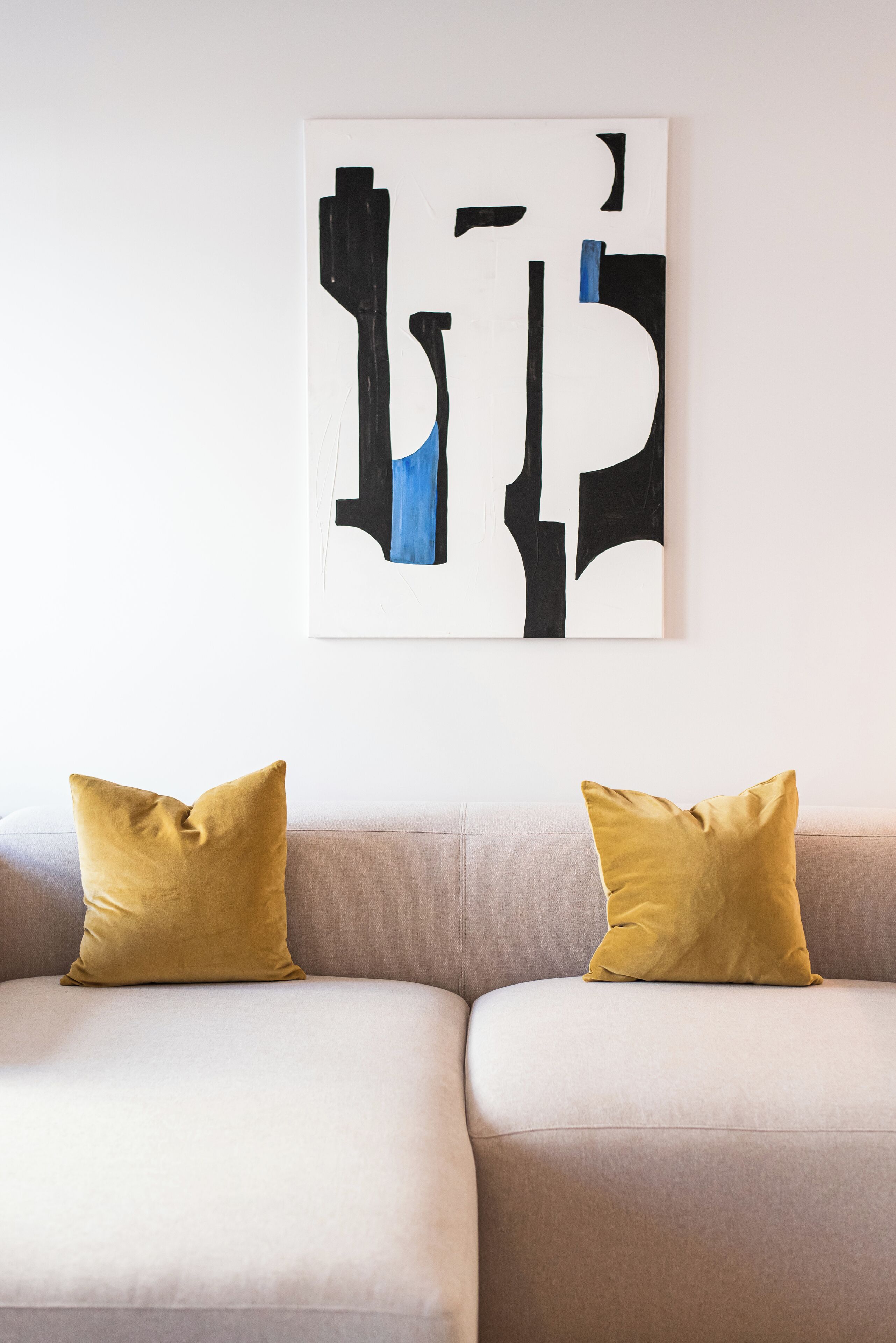 A modern living room with a beige sofa, two mustard-colored pillows, and a large abstract painting on the wall.