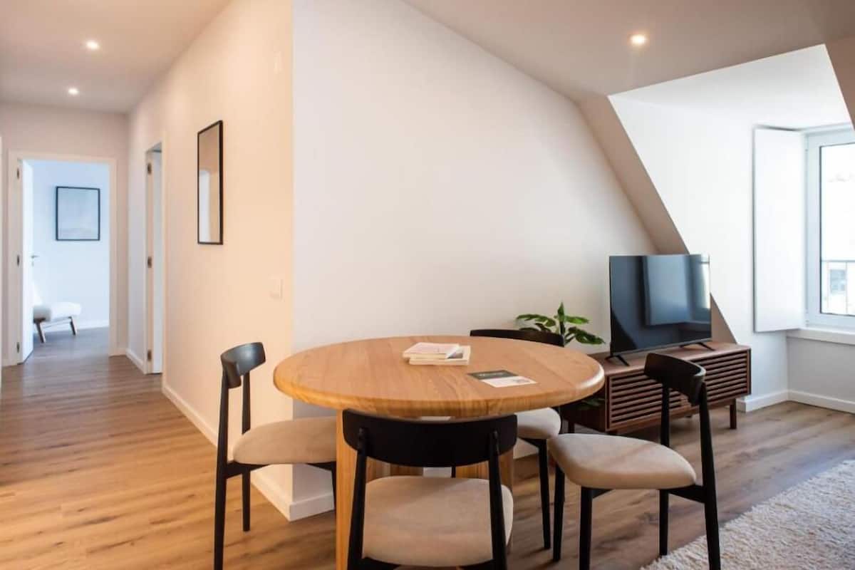 Una sala de estar moderna con una mesa redonda de madera, sillas negras, un televisor de pantalla plana y una planta.