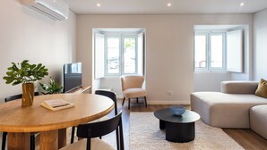 Een moderne woonkamer met een ronde houten tafel, een beige bank en een zwarte salontafel. Er is een witte airconditioningunit aan de muur, een raam met uitzicht en een plant in een vaas.