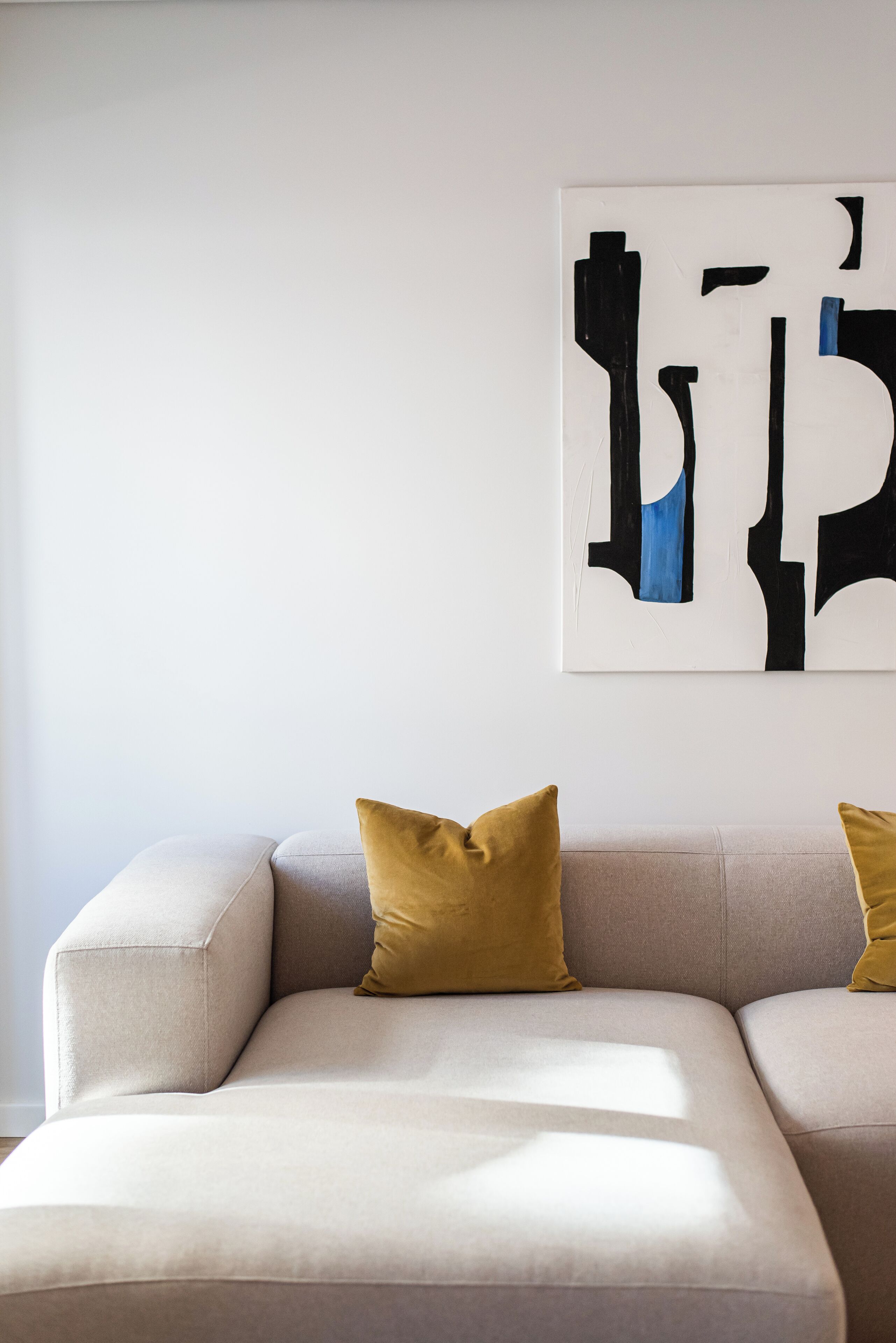 Een moderne woonkamer met een beige bank, twee mosterdgele kussens en een groot abstract schilderij aan de muur.