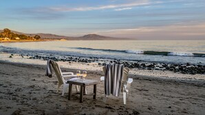 A beach with two chairs and a table, a glass of wine, and a view of the ocean and mountains. - The Cottage at Miramar (Montecito)