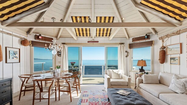 A spacious living area with a balcony overlooking the sea, featuring a dining table, sofa, and a patterned rug. - The Cottage at Miramar (Montecito)