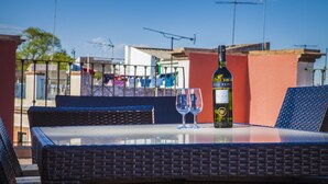 Una mesa con una botella de vino y dos copas en un balcón con mobiliario de mimbre y vistas a edificios.