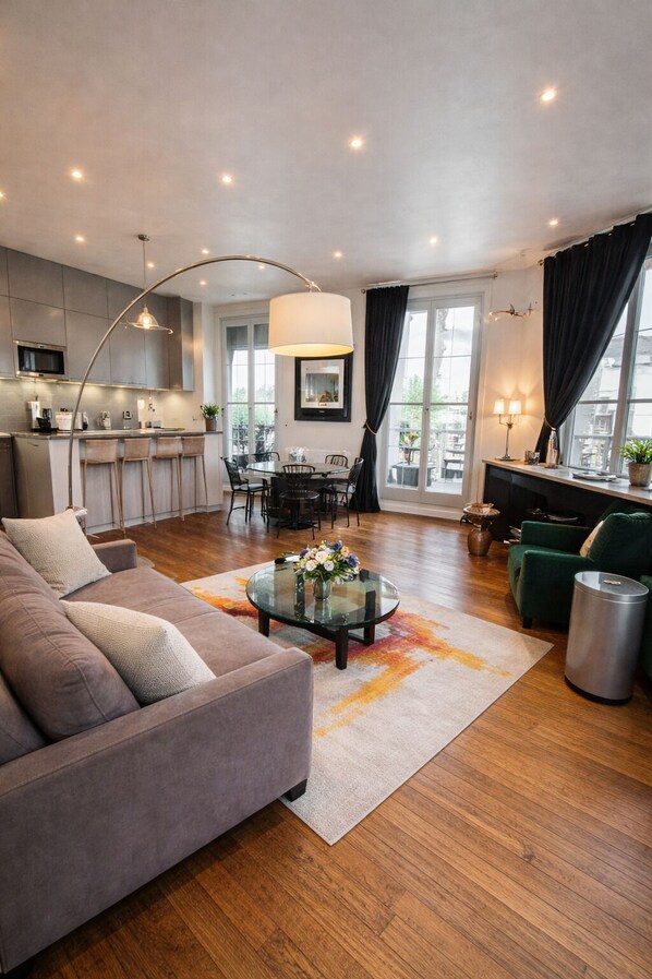 A modern living room with a sofa, a coffee table with flowers, a dining area with chairs, and a kitchen with a bar.