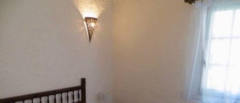 A bedroom with a single bed, a nightstand, a wall-mounted light fixture, and a window with curtains.