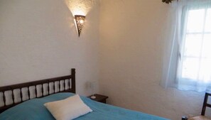Un dormitorio con una cama individual, una mesita de noche, una lámpara de pared y una ventana con cortinas.